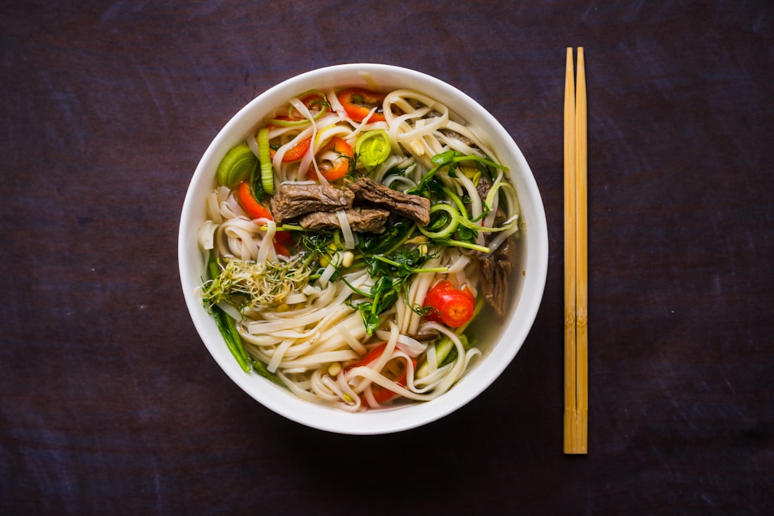 A bowl of noodle soup with chili oil blooming across the surface, herbs on the side